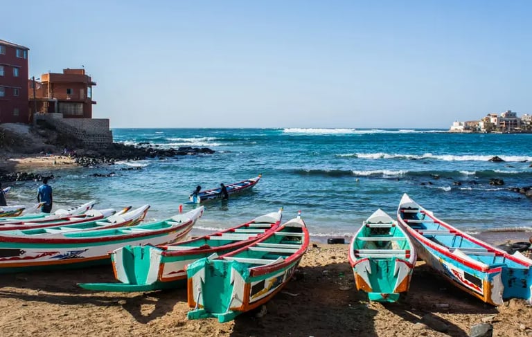 Colorful wooden fishing boats docked on a sandy beach in Senegal with ocean waves in the background.