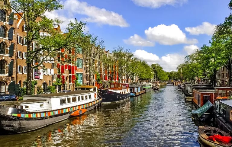 Traditional houseboats and brick buildings line a scenic Amsterdam canal under a blue sky.