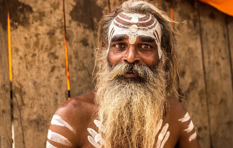A Hindu Sadhu with traditional white Tilak face paint and a long beard poses against a wooden background.