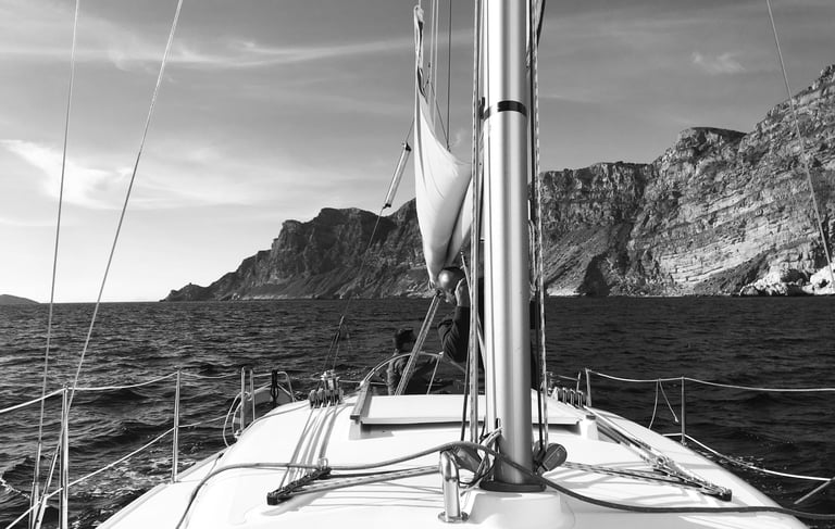 Black and white photo of the bow of a boat in the ocean in front of cliffs in good weather