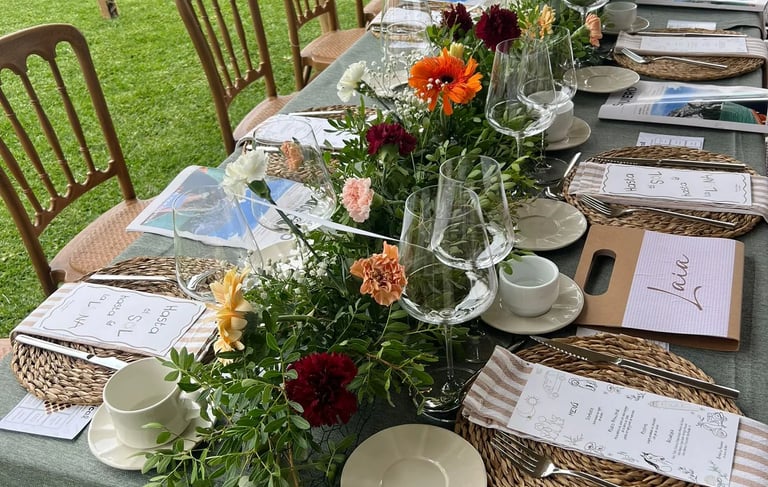 Long outdoor banquet table set with green linen, woven placemats, and a colorful floral centerpiece.