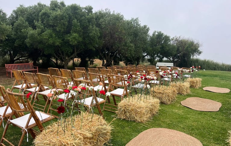Rustic wedding ceremony setup with bamboo chairs, straw bales with carnations, and woven jute rugs on the grass.