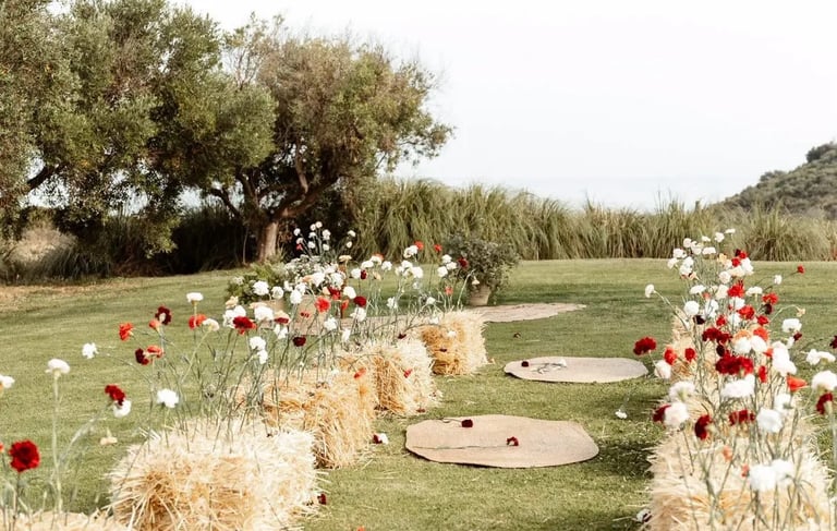 Rustic outdoor ceremony aisle with straw bales, red and white flowers, and woven jute rugs on a grassy lawn.