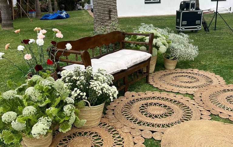 Rustic wooden bench with white cushion, surrounded by flowers and round woven jute rugs on a grassy lawn.