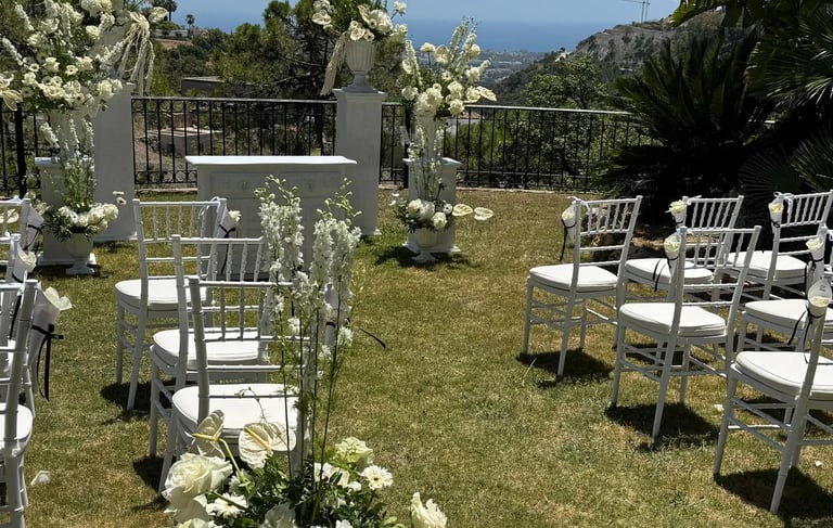 Outdoor wedding ceremony setup with white chairs, lush floral arrangements in white urns, and a scenic ocean view.