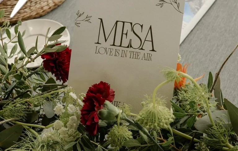 Elegant wedding table centerpiece with green foliage, red carnations, and a card reading 'MESA LOVE IS IN THE AIR'