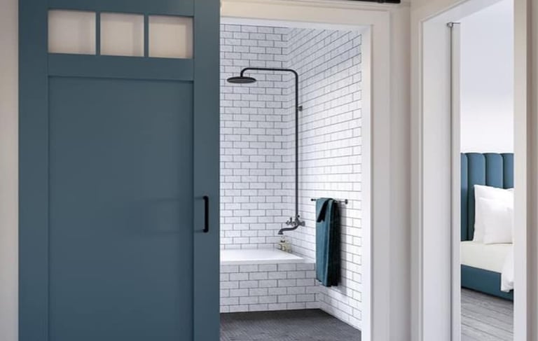 A modern blue sliding barn door with windows opening into a white subway tile bathroom with a black shower head.