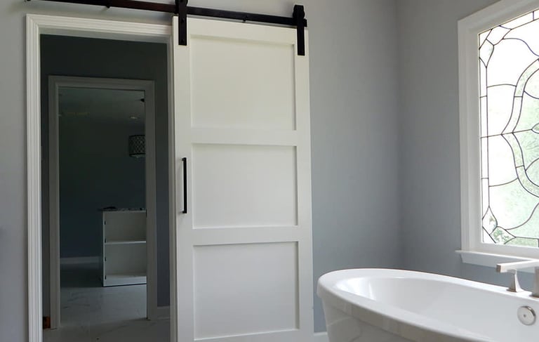 White 3-panel sliding barn door with black hardware installed in a modern gray bathroom.