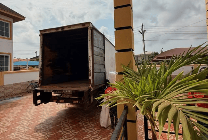 An open white moving truck with weathered sides and license plate visible, parked on a wavy red bric