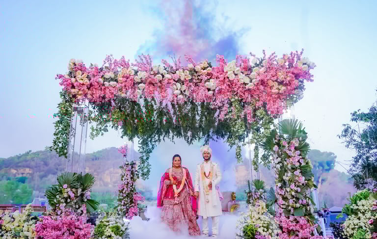 Indian bride and groom at an outdoor wedding ceremony under a floral mandap with colorful smoke.