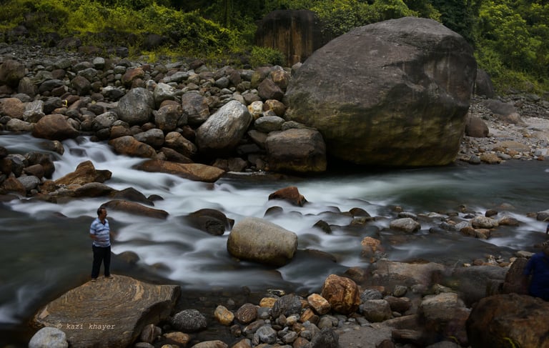 Picture of a river water caught in long exposure surrounded by big boulders on which there is a man.