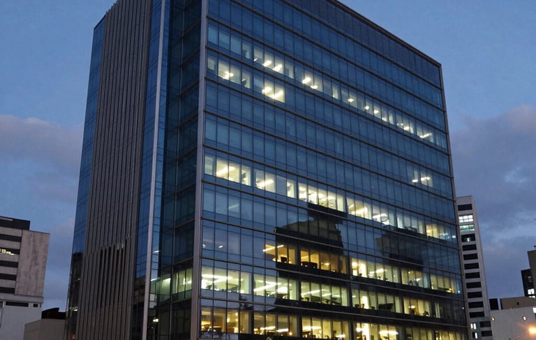 A wide photography shot of a modern corporate building in a South American / Brazilian city at dusk, with lights glowing behind large glass windows. The composition reflects expertise and reliability, using a palette of dark blue and steel blue. Soft architectural lighting creates a professional and efficient mood.