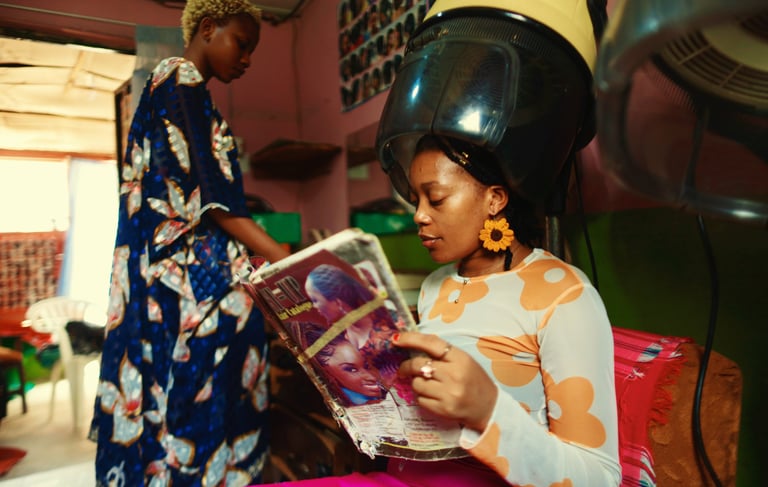 a woman in a colorful dress is reading a magazine while sitting under dryer in a hairdresser's shop 