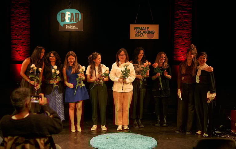 Group of women on stage holding flowers while receiving applause at the end of a storytelling event.
