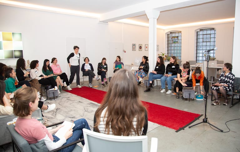 Woman walking down a red carpet toward a microphone, surrounded by seated women at a speaking event.