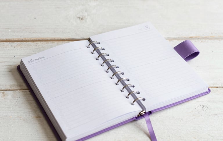 Flat lay of a beautifully crafted personalized creative planner and journal with lilac ribbons, lying on a clean whitewashed wooden surface in a Portuguese home design studio.