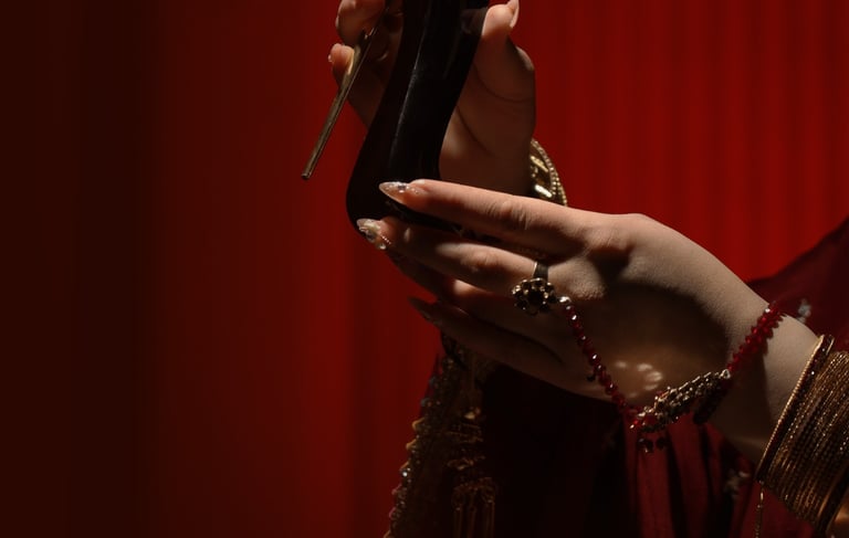 A hand with gold jewelry holding a black high-heel perfume bottle against a red background.