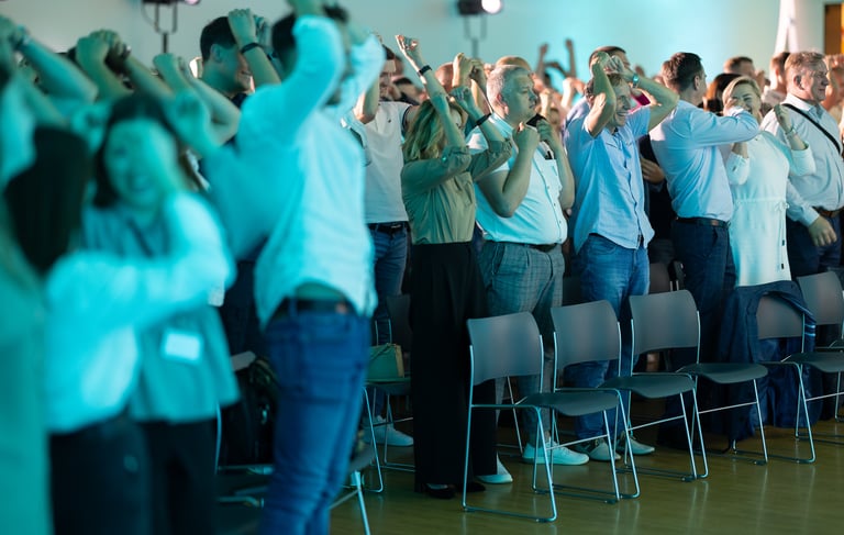 Nuotrauka iš įmonės renginio. Vyksta konferencija. Žmonės ploja atsistoję.