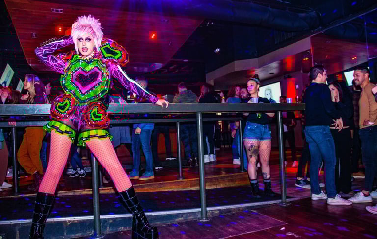 A drag queen in a neon punk outfit performs at a crowded nightclub with vibrant stage lighting.