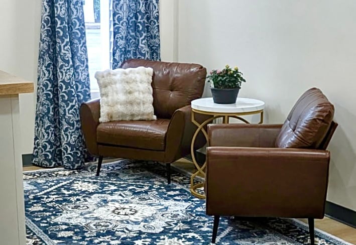 Two brown leather armchairs on a blue patterned rug with a white side table and damask curtains.