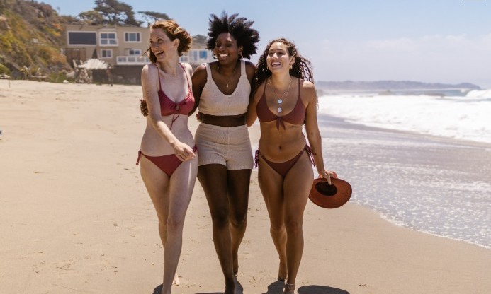 Three diverse women walking and laughing on a sunny beach in stylish bikinis and summer swimwear