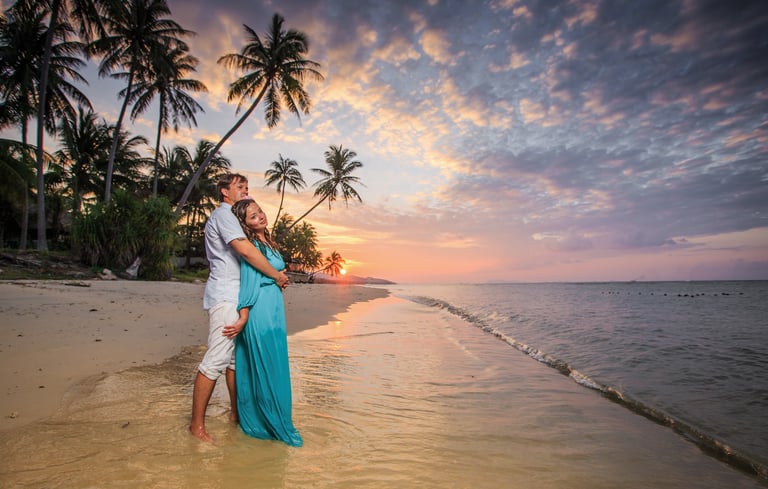 A couple embracing on a tropical beach at sunset, with palm trees and the ocean in the background