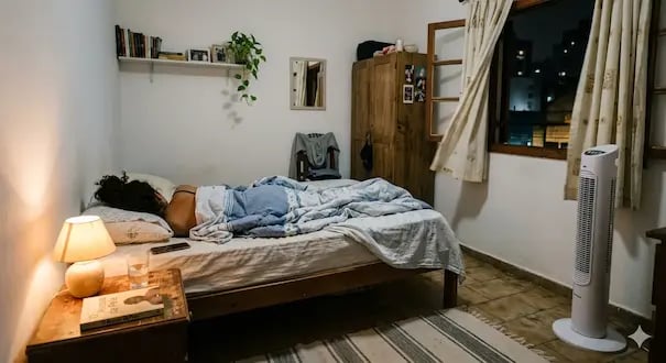 A person sleeping in a cozy bedroom with a warm bedside lamp and a tall white tower fan.