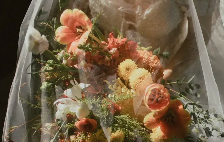 Novia con vestido de encaje bajo velo transparente sosteniendo un ramo en tonos naranja, blanco y verde.