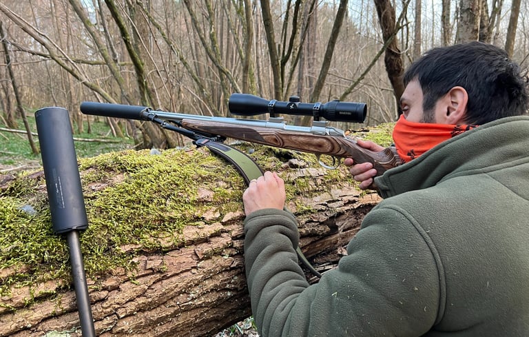 A stalker in a green fleece taking aim with a scoped rifle moderator rested on a moss-covered fallen