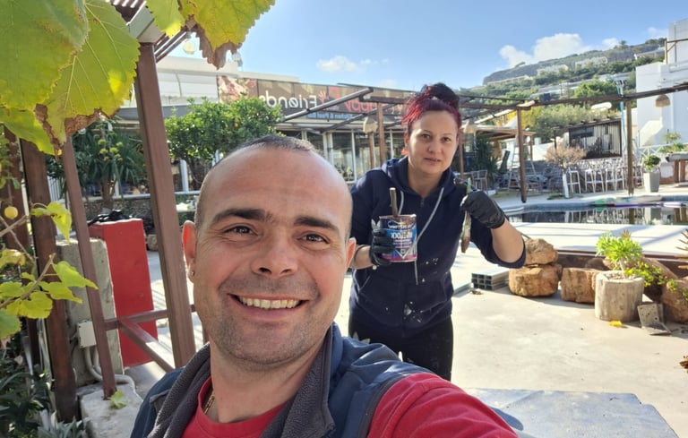 A smiling man and woman performing outdoor renovations near a swimming pool with a can of wood stain.