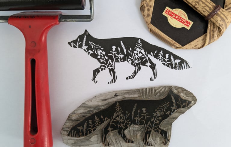 Handmade fox linocut print with floral silhouette detail, carving block, brayer, and baren tool.