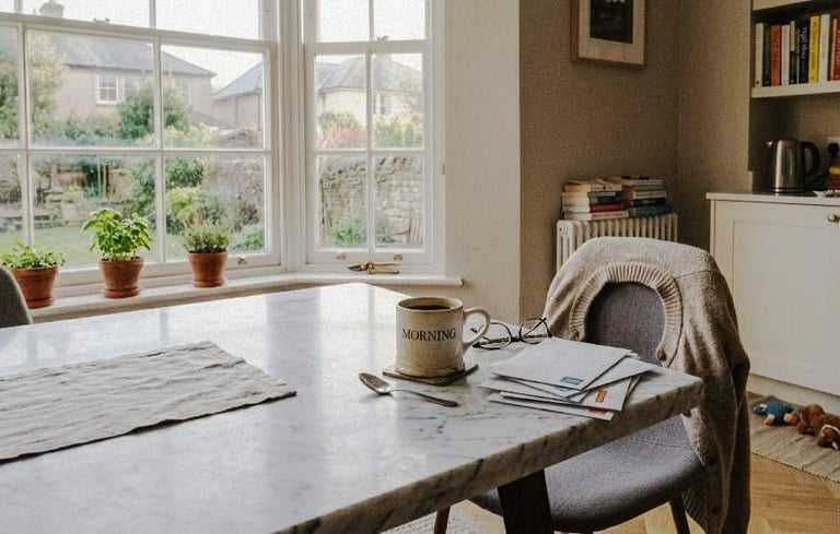 A kitchen table with a mug, scattered post, and a chair with a jumper in soft morning light