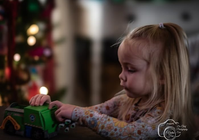 Toddler girl playing with a green toy truck near a blurred Christmas tree with lights. Athens, Oh