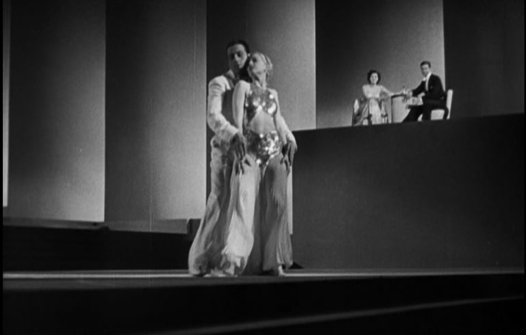Distant couple in a shadowy nightclub scene watching dancers perform in the moody foreground.