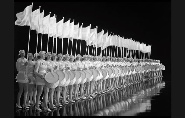 Two rows of chorus girls in short white military costumes with drums and flags in “All’s Fair in Lov