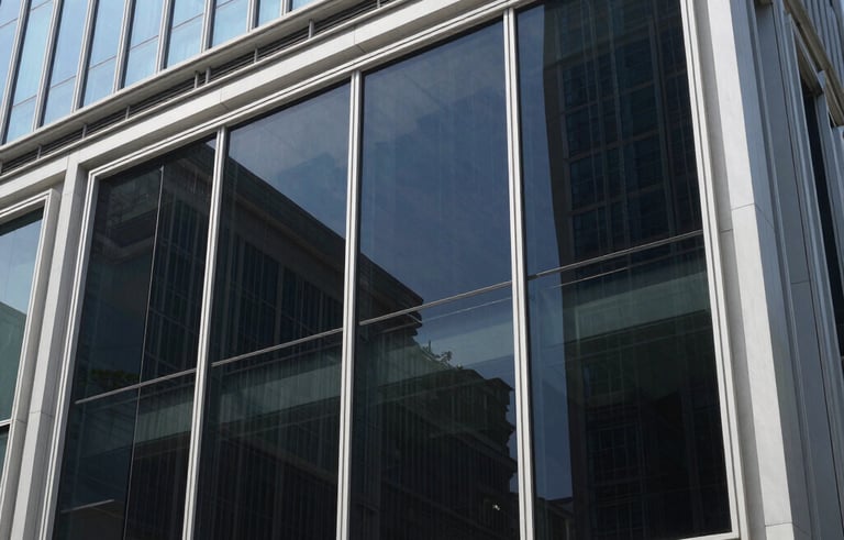 Photography of a modern glass building in an Indonesian city during a bright day, showing professional heat-rejecting window tinting being applied to large glass panels. The scene is clean, reflecting a blue sky, with a focus on the sleek dark tint finish.