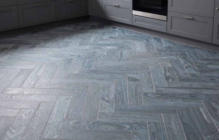 Wide shot of a modern North American kitchen featuring a stunning herringbone tile floor in muted blue-grey. The lighting is natural and soft, showcasing the precision of the installation and the sophisticated atmosphere of the home.
