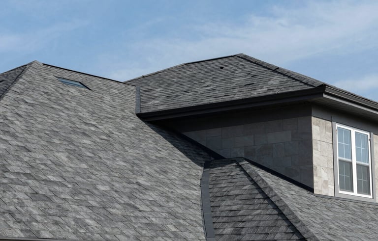 A low-angle, wide-shot photograph of a newly installed, modern roof on a large suburban home in North America, featuring clean lines and high-quality dark gray architectural shingles under a blue sky.