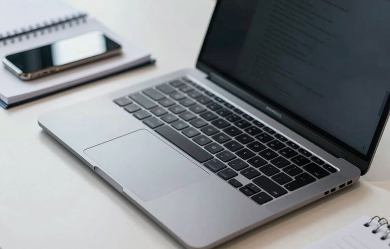 A close-up of a professional desk in an Indonesian office with a high-end laptop, a smartphone, and a notebook. The desk is organized and neat. The color scheme is steel blue and off-white, emphasizing a commitment to efficient technology and structured work systems.
