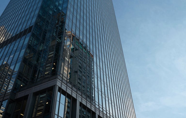 A low-angle shot of a modern glass skyscraper in a North American business district. The glass reflects a clear blue sky, with dark navy shadows and very light blue highlights, conveying a professional and high-trust atmosphere.