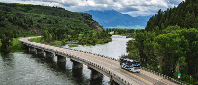 A luxury motorhome RV drives across a bridge over a river with mountains in the background.