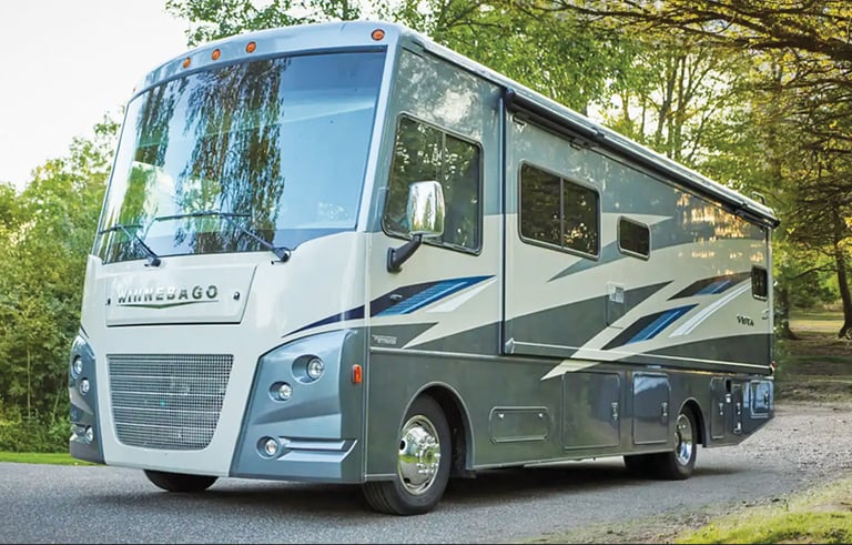 A Winnebago Vista Class A motorhome parked on a paved road surrounded by lush green trees.