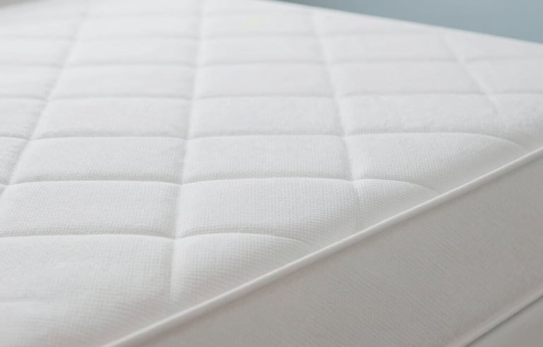 A professional close-up photograph of a bright, clean white mattress with crisp fabric texture. The setting is a minimalist bedroom with soft natural lighting and light blue accents. The image looks fresh, sterile, and inviting. South American home style.