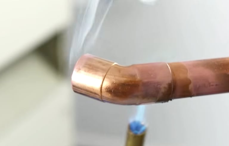 Professional plumber soldering a copper pipe elbow joint with a propane torch and visible smoke.