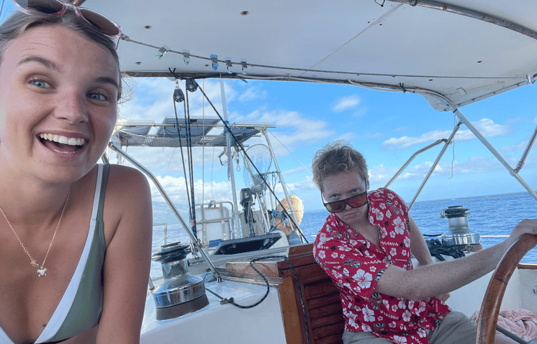A smiling woman and a man in a red floral shirt steering a sailboat on the open ocean.