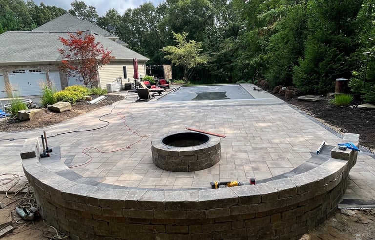 Custom paver patio installation with stone retaining wall, fire pit, and pool deck in a backyard.