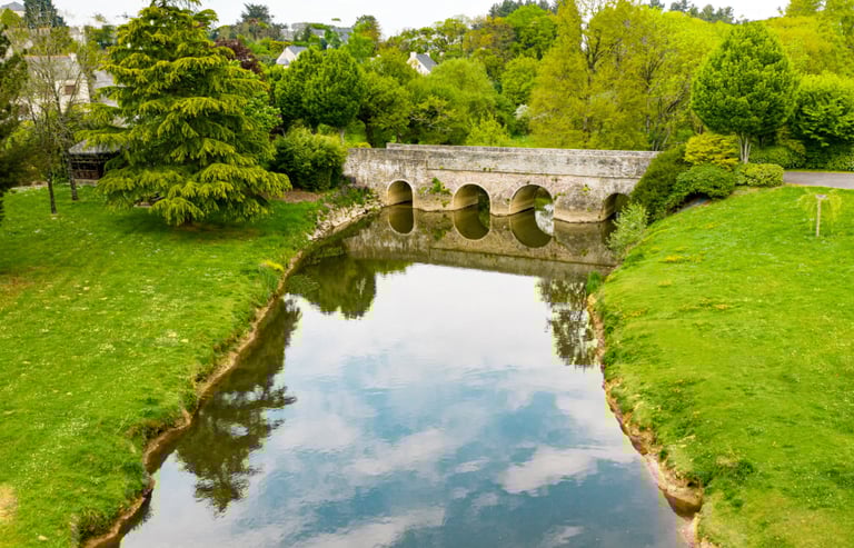 Pont historique en pierre se reflétant dans une rivière calme, entouré d'arbres et d'herbes verdoyan