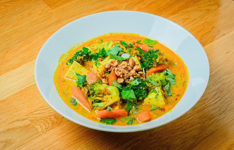Bowl of vietnamese curry with broccoli, carrots and herbs topped with crushed peanuts on a wooden t