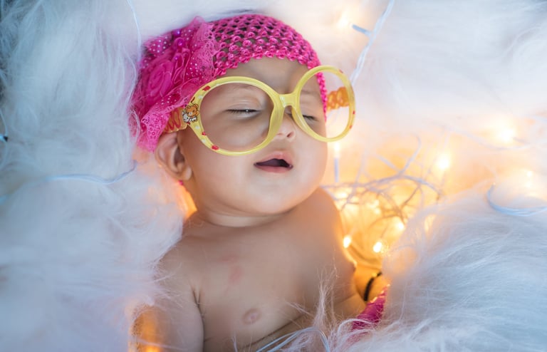 A newborn baby wearing oversized yellow glasses and a pink headband sleeps peacefully.
