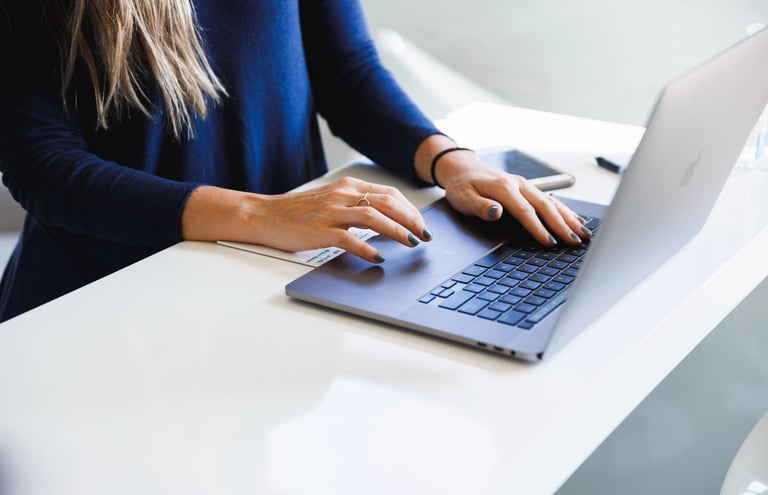 Femme habillée en bleu assise à son bureau blanc et qui pianote sur son ordinateur portable